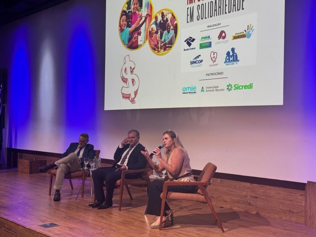 A imagem mostra uma apresentação em um auditório, com três palestrantes sentados em cadeiras na frente de uma tela. A tela exibe o título "Em Solidariedade", juntamente com os logotipos de diversas organizações envolvidas no evento, incluindo a Receita Federal, SINCOP, OMIE, Sicredi, e a Universidade Anhembi Morumbi. Há também imagens de pessoas, sugerindo um tema relacionado a ações sociais ou filantropia. Os palestrantes parecem estar envolvidos em uma discussão ou debate, e o ambiente é formal, típico de um evento corporativo ou acadêmico.