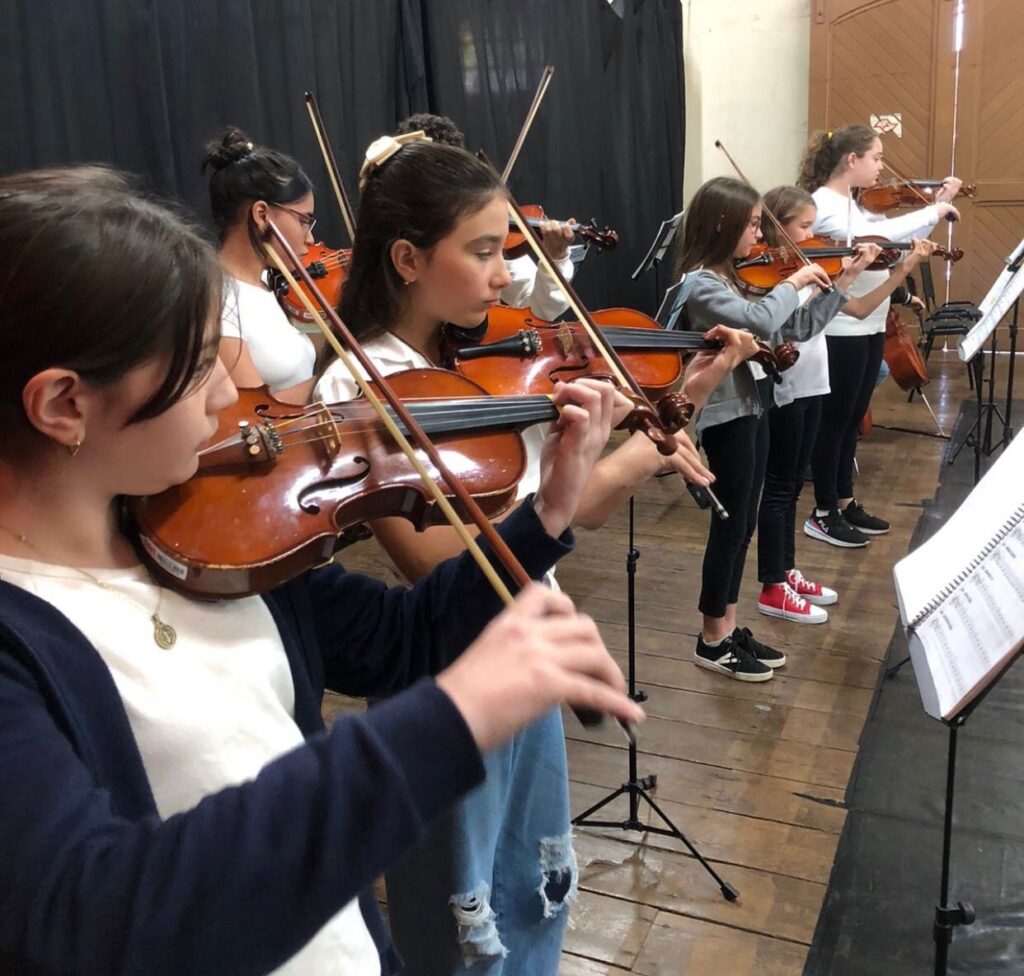 A imagem mostra um grupo de jovens tocando violino. Eles estão em uma sala com piso de madeira e um fundo escuro. A iluminação destaca os rostos concentrados dos músicos e os violinos que eles seguram. As partituras estão dispostas em estantes à frente deles. A cena transmite uma atmosfera de aprendizado e dedicação à música.