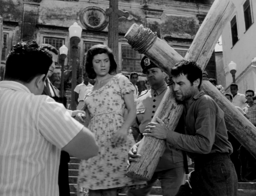 A imagem em preto e branco mostra uma cena movimentada em uma rua, possivelmente durante uma procissão religiosa. Um homem carrega uma pesada cruz de madeira nos ombros, enquanto uma mulher de vestido floral observa a cena com uma expressão preocupada. Ao redor, outras pessoas, incluindo soldados e espectadores, acompanham a procissão. O cenário é de uma cidade antiga, com prédios históricos ao fundo. A atmosfera é tensa e dramática, sugerindo um evento significativo e possivelmente religioso. A fotografia é de alta qualidade, com detalhes nítidos e contraste bem definido. A composição da imagem é dinâmica, com as linhas da cruz e as pessoas criando um movimento visual que conduz o olhar do espectador pela cena.