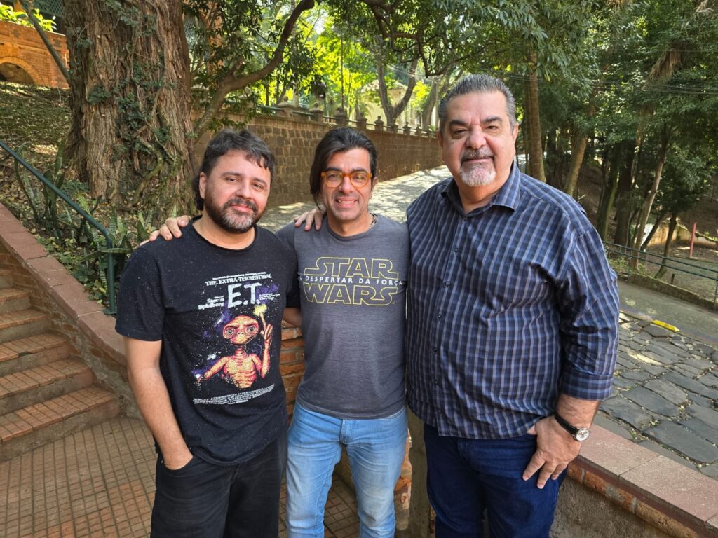 A imagem mostra três homens em pé juntos ao ar livre, em frente a uma parede de pedra e árvores frondosas. O homem da esquerda está usando uma camiseta preta com uma imagem de E.T. O homem do meio veste uma camiseta cinza com o logo de Star Wars. O homem da direita usa uma camisa xadrez azul e preta. Todos os três homens parecem estar sorrindo e se abraçando. O cenário é um ambiente externo, possivelmente um parque ou jardim, com uma escada de pedra visível ao fundo à esquerda. A iluminação sugere que a foto foi tirada durante o dia. A composição da foto é simples, com os três homens como foco principal, e o fundo desfocado, mas ainda visível. A imagem transmite uma sensação de camaradagem e amizade entre os três indivíduos.