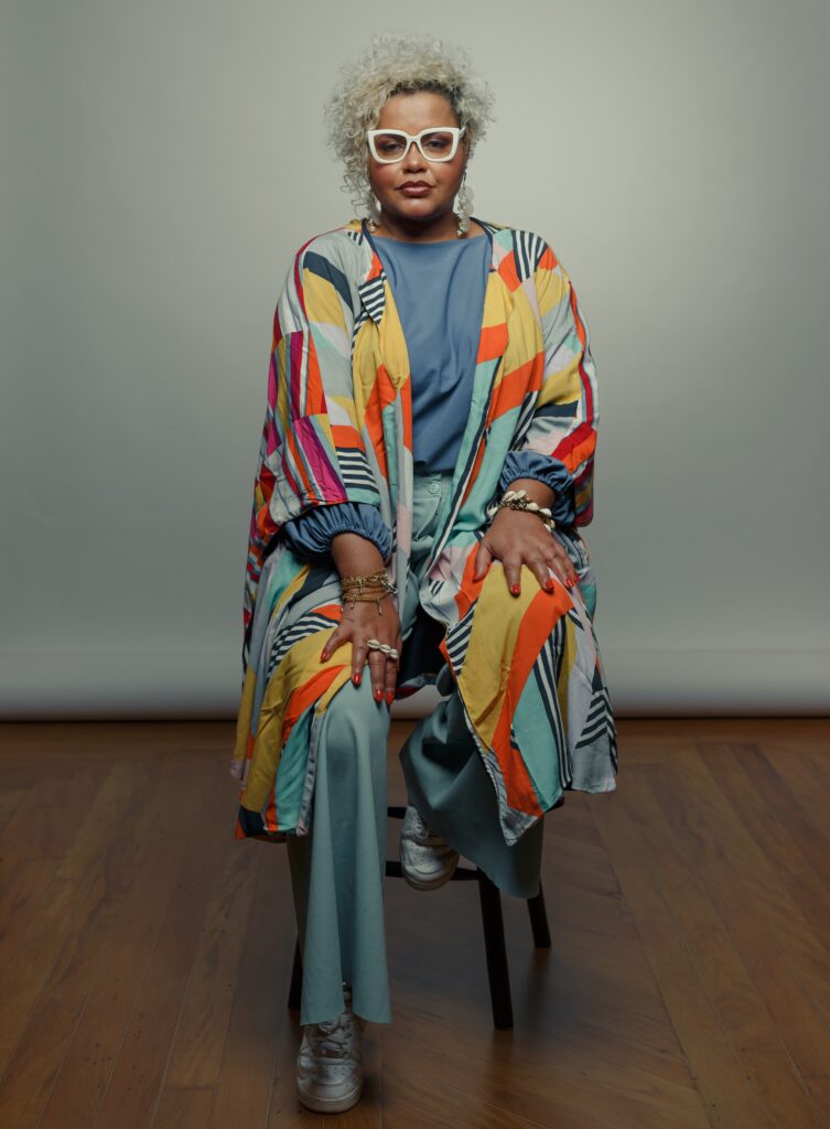 A imagem mostra uma mulher de meia-idade com cabelo curto e grisalho, usando óculos brancos de armação fina. Ela está sentada em um pequeno banco de madeira, vestindo uma calça comprida verde-azulada e uma quimono colorida com padrões geométricos em tons de laranja, amarelo, rosa, azul e branco. As mangas da quimono são compridas e largas, e ela está usando vários braceletes e anéis. Seus dedos têm unhas pintadas de vermelho. A expressão facial dela é séria e ela parece estar olhando diretamente para a câmera. O fundo é uma parede cinza clara e o chão é de madeira escura. A iluminação é suave e difusa. A imagem transmite uma sensação de serenidade e elegância.