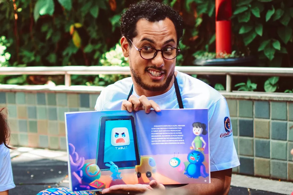 A imagem mostra um homem sorrindo enquanto segura um livro aberto para uma criança. O livro tem ilustrações coloridas de um tablet com um rosto zangado, um coelho azul, um carro de brinquedo e dois personagens semelhantes a insetos. O homem está usando uma camiseta branca com um logotipo no ombro. Ele tem cabelo cacheado escuro e usa óculos redondos. O fundo é desfocado, mas parece ser um ambiente externo com folhagem verde e uma parede de azulejos.