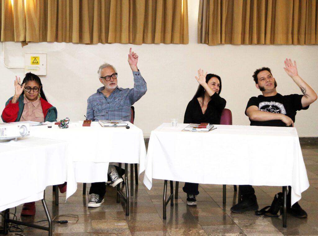 A imagem mostra quatro pessoas sentadas em frente a mesas cobertas com toalhas brancas, em uma sala com cortinas amarelas ao fundo. As quatro pessoas estão com as mãos levantadas, como se estivessem votando ou participando de uma discussão. A pessoa mais à esquerda é uma mulher com óculos e cabelos escuros, vestindo uma jaqueta verde e vermelha sobre uma camisa. A segunda pessoa, um homem mais velho com óculos e barba grisalha, veste uma camisa xadrez azul. A terceira pessoa é uma mulher com cabelos escuros, usando uma blusa preta. A pessoa mais à direita é um homem com cabelo escuro e costeletas proeminentes, vestindo uma camiseta preta com uma estampa. Na mesa à frente da primeira pessoa, há um projetor. Nas mesas, também há papéis, canetas e copos. O piso é de ladrilhos com um padrão irregular. Ao fundo, há uma caixa elétrica com um símbolo de aviso de perigo.