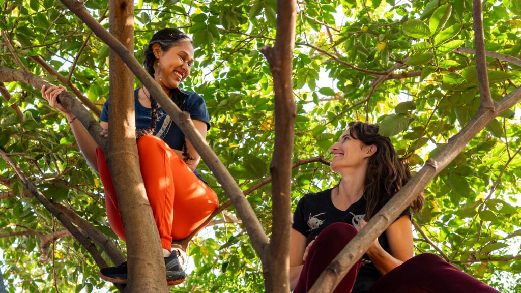 A imagem mostra duas mulheres sorrindo, sentadas em uma árvore frondosa. A mulher à esquerda está usando calças laranja e uma camiseta azul-marinho, enquanto a mulher à direita está usando calças vinho e uma camiseta preta. Ambas as mulheres parecem estar se divertindo e desfrutando da companhia uma da outra. A árvore é alta e cheia de folhas verdes vibrantes, sugerindo um ambiente tropical ou subtropical. A luz do sol ilumina as folhas, criando um efeito brilhante e arejado. O fundo é desfocado, mas mostra mais folhas e galhos, reforçando a sensação de estar dentro de uma floresta ou jardim exuberante. As mulheres estão posicionadas de forma que seus rostos estejam voltados uma para a outra, sugerindo uma conversa ou interação íntima. A imagem transmite uma sensação de alegria, amizade e conexão com a natureza. A composição da imagem é dinâmica, com as mulheres posicionadas de forma diagonal e os galhos da árvore criando linhas que levam o olhar do observador para as mulheres. A fotografia é bem iluminada e nítida, mostrando detalhes tanto das mulheres como das folhas da árvore.