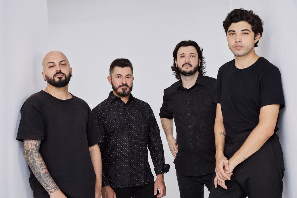 A imagem mostra quatro homens em pé contra uma parede branca. Todos estão vestidos de preto. O homem mais à esquerda é careca e tem tatuagens no braço. Ele está usando uma camiseta preta. O homem ao seu lado tem cabelo curto e escuro e está usando uma camisa social preta com pequenos pontos brancos. O terceiro homem tem cabelo comprido e escuro e está usando uma camisa social preta com um padrão sutil. O homem mais à direita tem cabelo curto e escuro e está usando uma camiseta preta. Todos os homens parecem estar na faixa dos 20 ou 30 anos. Eles parecem estar posando para uma foto. O fundo é branco e simples. A iluminação é suave e uniforme. A imagem é nítida e bem composta.