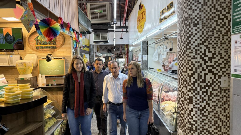 A imagem mostra um grupo de pessoas caminhando por um corredor em um mercado. No lado esquerdo, há uma barraca com produtos alimentícios, incluindo recipientes de comida amarela em uma vitrine refrigerada e um dispensador de suco de milho. Acima da barraca, há decorações coloridas, incluindo um leque de papel arco-íris e bandeirolas. No lado direito, há uma vitrine refrigerada cheia de carne crua, com um açougueiro trabalhando ao fundo. Um pilar com um padrão de mosaico marrom e branco se destaca na frente da vitrine de carne. Há sinais e iluminação típicos de um mercado.