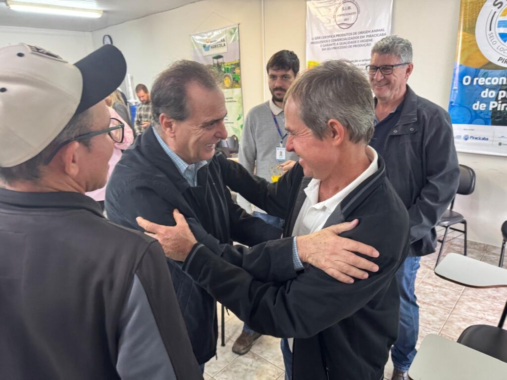 A imagem mostra um grupo de homens em um evento. Dois homens no centro estão se cumprimentando com um abraço, ambos sorrindo. O homem à esquerda usa uma jaqueta preta sobre uma camisa xadrez azul e branca. O homem à direita usa uma jaqueta preta sobre uma camisa branca. Ao fundo, há um banner com o texto "SIM CERTIFICA PRODUTOS DE ORIGEM ANIMAL" e "PIRACICABA", sugerindo que o evento está relacionado à agricultura ou produção de alimentos na região de Piracicaba. Há também outros homens em segundo plano, um deles usando um boné e óculos. As paredes parecem ser de uma sala de reuniões ou auditório, com cadeiras e mesas visíveis.