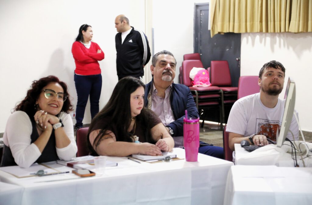 No primeiro plano, há três pessoas sentadas em uma mesa coberta com toalhas brancas. À esquerda, uma mulher com cabelos escuros e óculos está com as mãos apoiadas no queixo, sorrindo. Ao lado dela, uma mulher com cabelos longos e escuros está olhando para a frente, com as mãos sobre alguns papéis. Na frente dela, há um copo rosa com um canudo. À direita, um homem com barba está sentado em frente a um computador, olhando para cima. Ele usa uma camiseta branca com uma estampa. Ao fundo, em pé, há um homem e uma mulher. A mulher, vestindo uma blusa vermelha e calça azul, está de braços cruzados e conversando com o homem, que veste uma jaqueta preta com listras brancas e uma blusa branca por baixo. Atrás deles, há cadeiras vermelhas empilhadas e uma cortina amarela ao fundo.