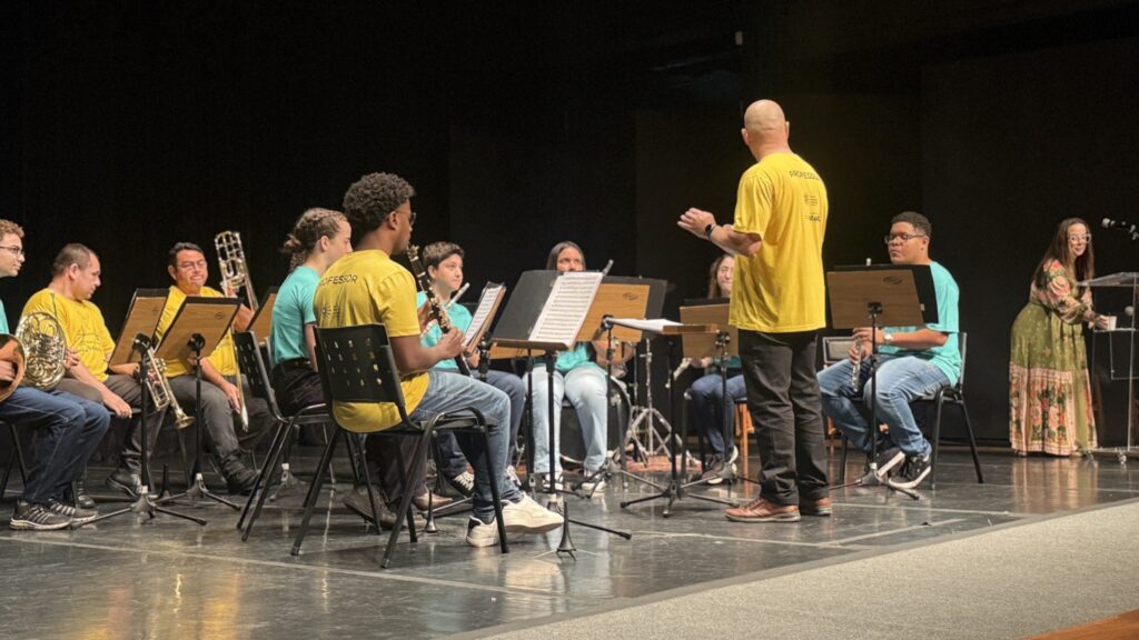 A imagem mostra uma banda de música em um palco, aparentemente durante um concerto ou ensaio. Há vários músicos sentados em cadeiras, cada um com seu instrumento. Os instrumentos incluem trompa, trompete, clarinete, flauta e talvez outros instrumentos de sopro. Os músicos usam roupas casuais, principalmente em tons de amarelo e azul-turquesa. Um maestro, vestindo uma camiseta amarela com a palavra "Professor" escrita, está de pé, conduzindo a orquestra. Ao fundo, uma mulher está em pé perto de um púlpito, possivelmente a regente ou uma pessoa envolvida na organização do evento. O palco é escuro, com iluminação focada nos músicos. A atmosfera parece calma e concentrada, sugerindo um ambiente profissional e organizado.