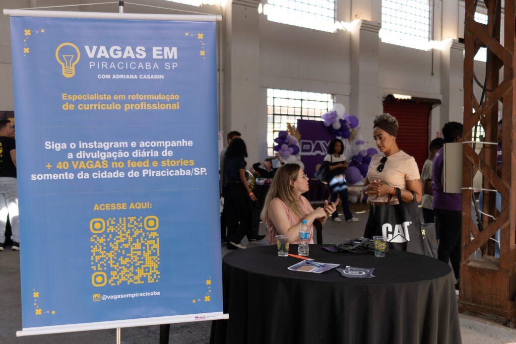 A imagem mostra um estande de recrutamento em um evento em Piracicaba, SP. Um banner azul claro anuncia vagas de emprego, com um QR code para acesso a mais informações e o Instagram @vagasempiracicaba. Duas mulheres estão conversando perto do banner, uma delas aparentemente uma recrutadora, e outras pessoas circulam pelo local, que parece ser um galpão industrial adaptado para o evento. Há balões roxos e uma decoração discreta no fundo. O ambiente é informal e movimentado.