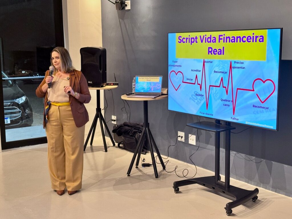 A imagem mostra uma mulher em pé, segurando um microfone e falando para uma plateia. Ela está em uma sala com uma tela grande atrás dela, exibindo uma apresentação sobre "Script Vida Financeira Real". A apresentação inclui um gráfico semelhante a um eletrocardiograma, com palavras como "Iniciar", "Acreditar", "Recomeçar", "Falhar" e "Aposentar" dispostas ao longo da linha. Há também um laptop em uma mesa ao lado da mulher, exibindo o mesmo título da apresentação. A sala parece ser um espaço moderno, com paredes cinzas e iluminação suave. Há caixas de som e outros equipamentos de apresentação visíveis. No geral, a imagem retrata uma palestra ou workshop sobre finanças pessoais.