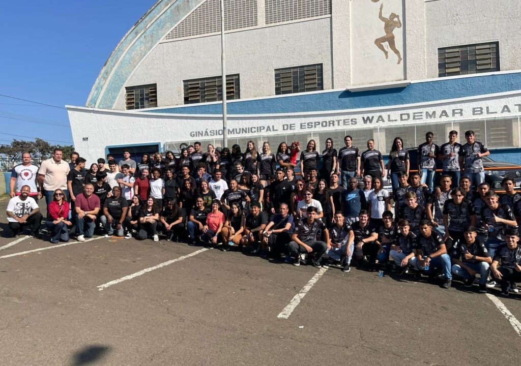 A imagem mostra um grande grupo de pessoas, predominantemente jovens, posadas para uma foto em frente a um ginásio municipal de esportes. Há mais de 50 indivíduos na foto, muitos vestindo roupas escuras, algumas com logotipos visíveis. Eles estão organizados em várias fileiras, com aqueles na frente sentados e os de trás em pé. Algumas pessoas na parte de trás parecem estar usando uniformes esportivos. O ginásio ao fundo é um edifício de estrutura simples, de cor branca e azul, com o nome "Ginásio Municipal de Esportes Waldemar Blat" visível na fachada. O dia parece ensolarado, com um céu azul claro visível atrás do grupo. O asfalto do estacionamento onde estão posicionados é escuro e apresenta marcações de vagas de estacionamento. Há também alguns carros visíveis ao fundo, à direita do grupo. A atmosfera geral sugere um evento esportivo ou relacionado à comunidade.