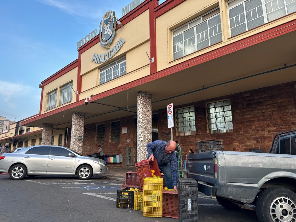 A imagem retrata uma cena em frente a um edifício com uma fachada amarela e detalhes em vermelho, possivelmente um mercado municipal, como indicado pela inscrição "MERCADO PIRACICABA" e o brasão no topo. O céu está azul com algumas nuvens. No primeiro plano, um homem vestindo uma camisa xadrez azul e jeans está agachado, manuseando caixas de frutas, aparentemente laranjas. As caixas são de plástico, algumas amarelas e outras vermelhas, e estão empilhadas no chão de asfalto. Há também caixas pretas. À esquerda, um carro prateado está estacionado. Ao fundo, outras pessoas podem ser vistas, algumas sentadas e outras em pé, e um caminhão cinza está estacionado à direita. O edifício possui janelas com grades e uma entrada coberta por um toldo, sustentado por colunas com um revestimento texturizado em tons de bege e marrom. Há também um sinal de trânsito com a letra "E" dentro de um círculo vermelho. A cena sugere uma atividade de carga ou descarga de mercadorias em um ambiente urbano.
