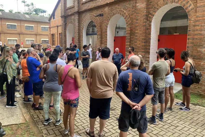 A imagem mostra um grupo de pessoas em frente a um edifício de tijolos com arcos. A maioria das pessoas está de pé e parece estar ouvindo alguém falar, possivelmente um guia turístico. O edifício tem portas vermelhas e uma arquitetura mais antiga. O tempo parece nublado.