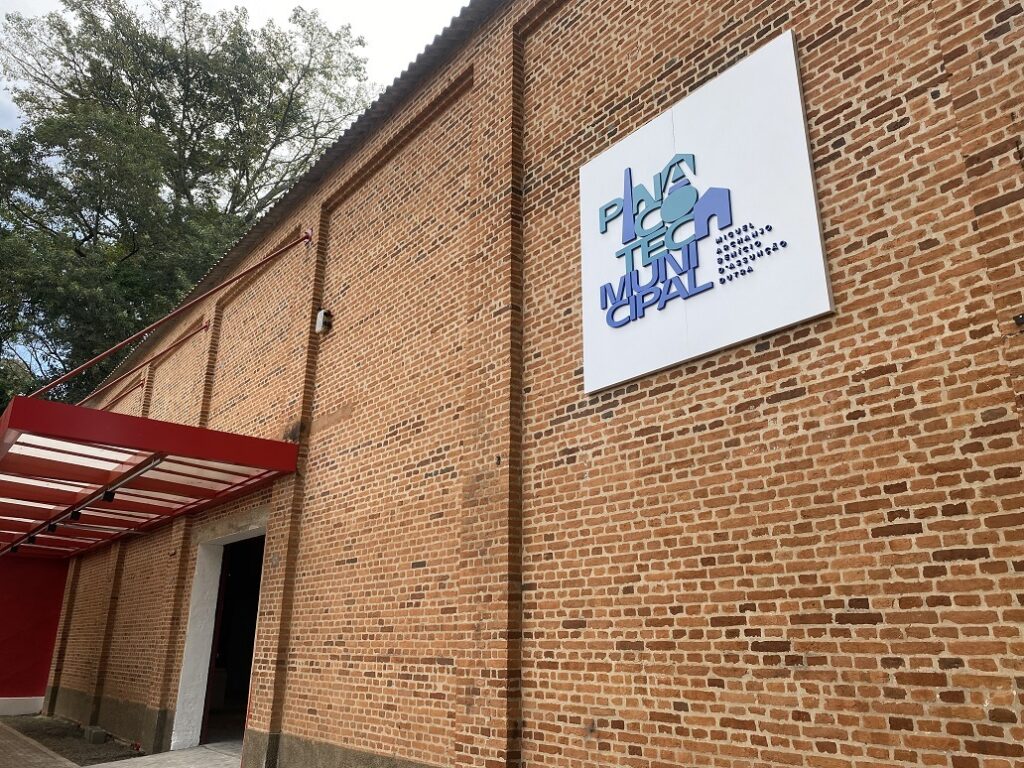 A imagem mostra um edifício de tijolo vermelho de um andar, com uma placa branca na parede externa que indica "Pinacoteca Municipal". A placa também contém informações adicionais em letras menores, possivelmente sobre o nome de um projeto ou organização. A arquitetura do edifício é simples e funcional, com uma entrada coberta por uma estrutura de metal vermelha. Há uma árvore verde visível ao fundo, sugerindo um ambiente arborizado. A iluminação e a qualidade da imagem sugerem que a foto foi tirada durante o dia, em condições de luz natural. Os tijolos são uniformes em cor e textura, e o edifício parece bem conservado. O estilo arquitetônico é bastante simples e não exibe detalhes ornamentais elaborados. A cor vermelha da cobertura contrasta com o tijolo vermelho-alaranjado do edifício, criando um elemento visual interessante. A imagem é nítida e bem iluminada, permitindo uma visualização clara de todos os detalhes do edifício e da placa.