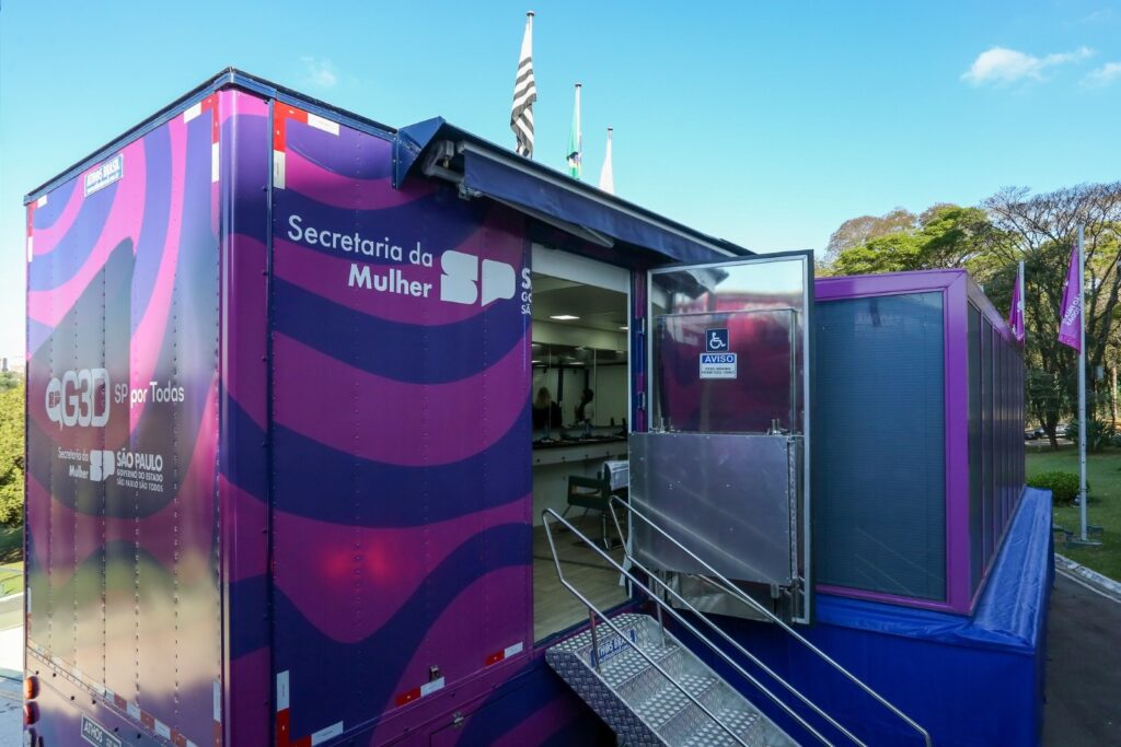 A imagem mostra um caminhão adaptado que serve como unidade móvel da Secretaria da Mulher do Estado de São Paulo. O caminhão é de cor roxa e possui a logo da secretaria, bem como a frase "SP por Todas". Ele está estacionado em um local com árvores e grama ao fundo. O caminhão possui uma rampa de acesso para pessoas com deficiência e uma porta lateral que dá acesso ao interior. É possível ver no interior alguns equipamentos e mobiliário, sugerindo um espaço para atendimento ao público. A parte traseira do caminhão é fechada, enquanto a lateral possui uma estrutura adicional, provavelmente para aumentar o espaço interno. A imagem sugere que o caminhão é utilizado para levar os serviços da Secretaria da Mulher para diferentes locais, facilitando o acesso da população a esses serviços. As bandeiras brasileiras e do estado de São Paulo no topo do caminhão reforçam a sua ligação com o governo estadual.