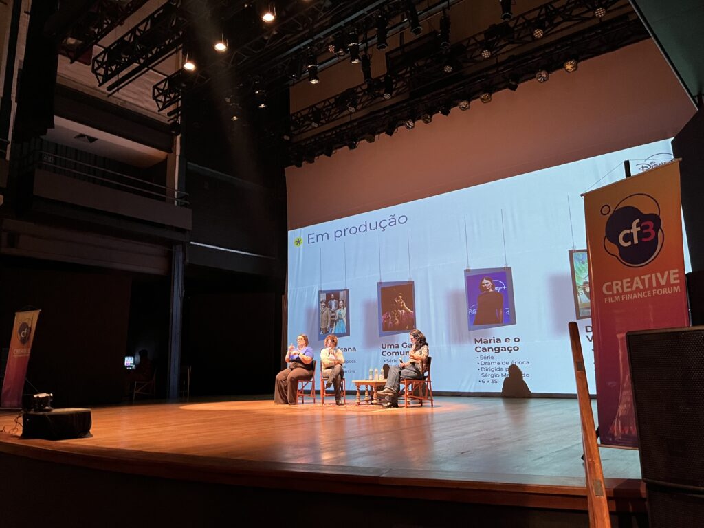 A imagem mostra um palco de teatro com três pessoas sentadas em cadeiras, aparentemente numa sessão de perguntas e respostas ou painel de discussão. Ao fundo, há uma tela grande exibindo o título "Em produção" e imagens promocionais de diferentes séries ou filmes, incluindo uma intitulada "Maria e o Cangaço". Há também um banner do "Creative Film Finance Forum", sugerindo que o evento é um fórum de financiamento para filmes. O ambiente é escuro, com iluminação focada no palco, indicando uma apresentação formal. O palco é de madeira polida e o local parece ser um auditório ou teatro. A atmosfera é profissional e focada na indústria cinematográfica.