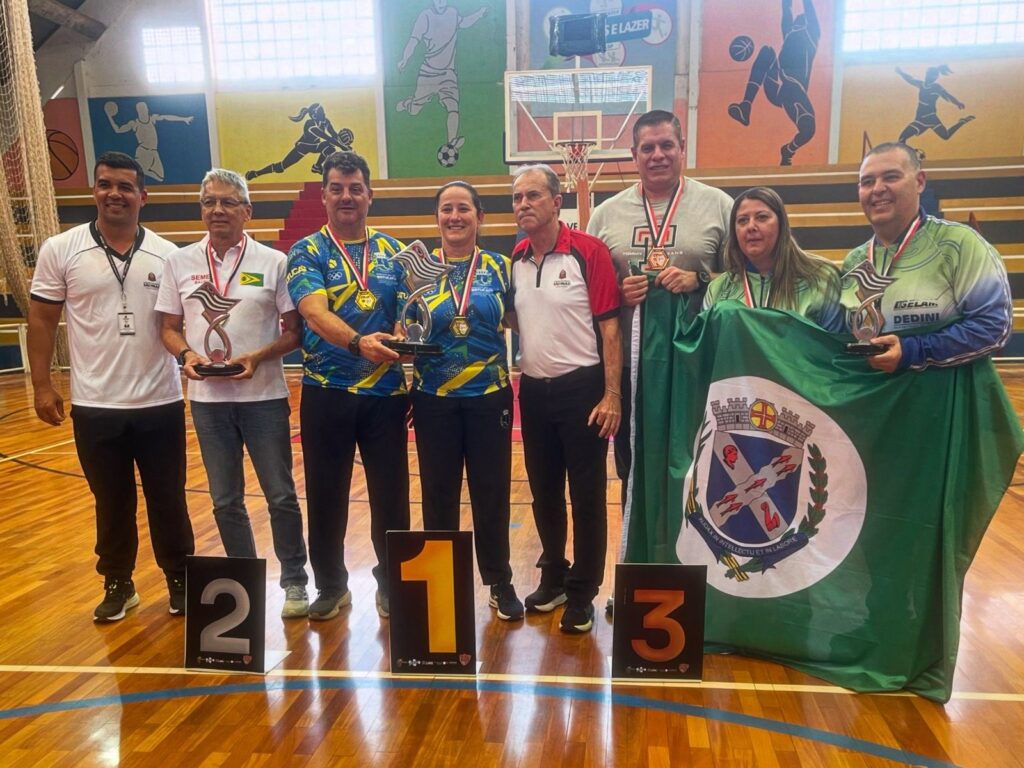 A imagem mostra um grupo de oito pessoas em uma quadra esportiva, possivelmente após uma competição. Todos estão sorrindo e posando para a foto. Seis pessoas estão de pé, enquanto duas estão sentadas em degraus ao fundo. No primeiro plano, há três placas pretas com números: "2", "1" e "3", indicando a classificação dos vencedores. As pessoas em primeiro, segundo e terceiro lugar estão vestindo medalhas e segurando troféus. As pessoas no pódio parecem ser: * *Primeiro lugar:* Um homem e uma mulher, ambos vestindo camisetas azuis e amarelas com medalhas de ouro. Eles seguram um grande troféu prateado. * *Segundo lugar:* Um homem mais velho, vestindo uma camiseta branca com uma medalha. Ele segura um troféu menor. * *Terceiro lugar:* Uma mulher, vestindo uma camiseta verde e uma medalha. Ela segura um troféu prateado. Ao fundo, há pinturas nas paredes retratando cenas esportivas, como futebol, basquete e atletismo. Há também uma quadra de basquete visível. A atmosfera geral é de celebração e conquista.