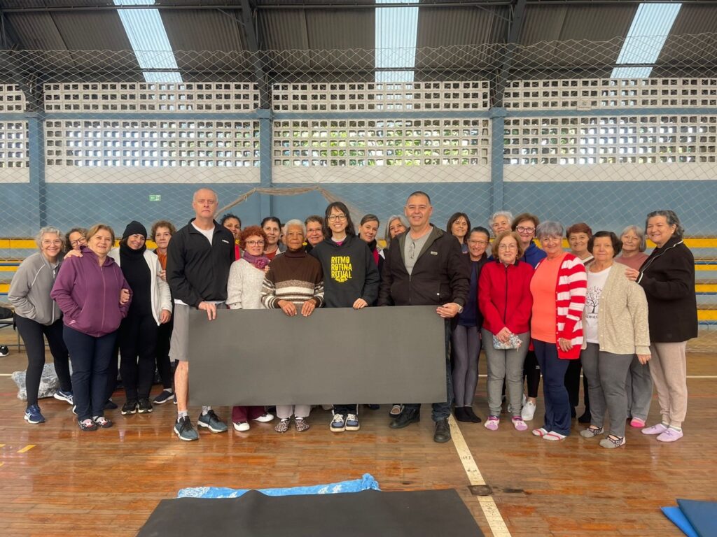 A imagem mostra um grupo de pessoas, a maioria mulheres idosas, em uma quadra poliesportiva, possivelmente após uma aula de ginástica. Elas estão sorrindo e posando para a foto, segurando um grande tapete cinza na frente delas. As pessoas vestem roupas esportivas e casuais. Há dois homens no grupo, um em pé no centro e outro à direita. O fundo da imagem mostra a estrutura da quadra, com grades de proteção e janelas altas. O piso é de madeira, típico de quadras esportivas. A iluminação parece ser artificial, vindo do teto da quadra.