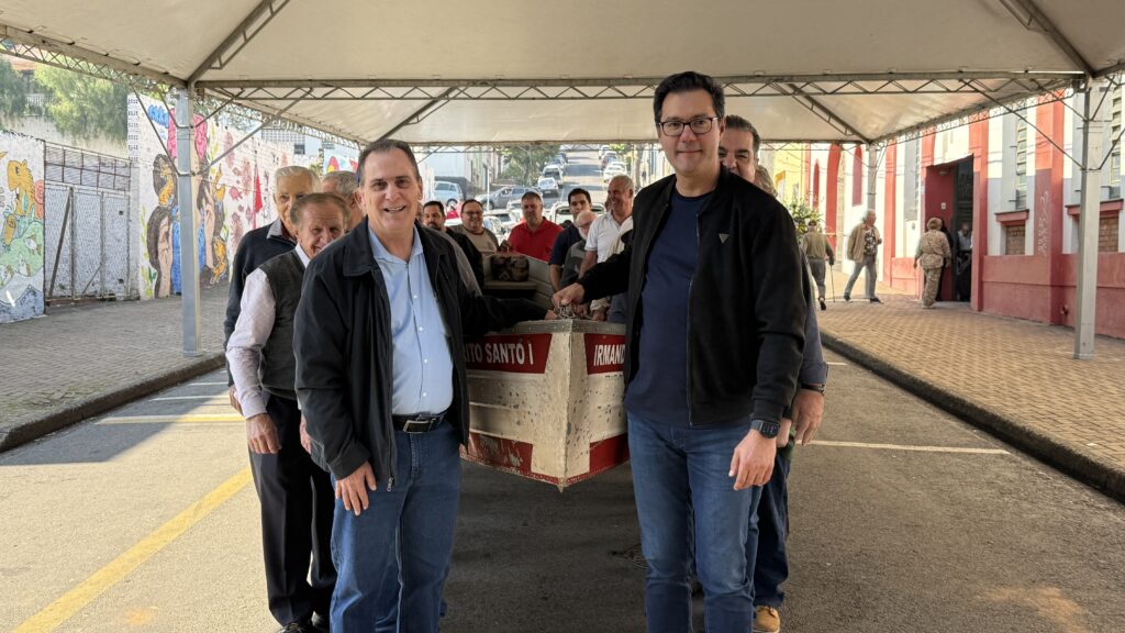 A imagem mostra um grupo de homens carregando um barco pequeno sob uma estrutura de lona. O barco parece ser de alumínio, com a inscrição "Santo I" visível. Os homens parecem estar sorrindo e parece haver um clima festivo ou comunitário. Há um fundo de rua com prédios e grafites. No total, parece ser uma cena de um evento comunitário ou uma celebração local. As roupas dos homens sugerem um ambiente casual e informal. A iluminação indica que a foto foi tirada durante o dia.