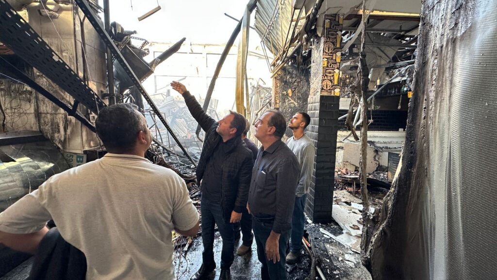 A imagem mostra um grupo de homens observando os estragos causados por um incêndio em um mercado ou centro comercial. O local está severamente danificado, com estruturas metálicas retorcidas, paredes queimadas e destroços espalhados