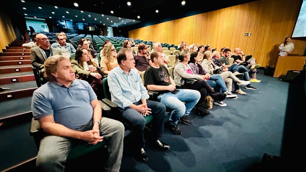 A imagem mostra uma plateia sentada em cadeiras em uma sala de conferências. A maioria das pessoas está olhando para a frente, em direção a um palco ou tela. A sala parece ter um design moderno, com assentos em tons de verde e azul, e paredes de madeira clara. Há uma pessoa em pé no palco, aparentemente apresentando algo. A iluminação é suave, criando uma atmosfera propícia para uma apresentação ou palestra. A plateia é composta por pessoas de diferentes idades e gêneros, vestidas casualmente.