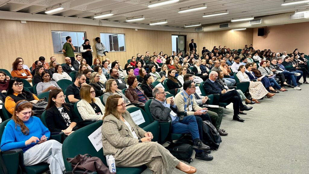 A imagem mostra uma sala de conferências cheia de pessoas sentadas em cadeiras, assistindo a uma apresentação. A maioria das pessoas está olhando para a frente, em direção a um palco ou tela que não está visível na imagem. Há uma mistura de homens e mulheres de várias idades e etnias. A sala parece ser moderna, com paredes de madeira e iluminação fluorescente. Algumas pessoas estão usando crachás, indicando que estão participando de um evento ou conferência. O ambiente geral é de atenção e engajamento.