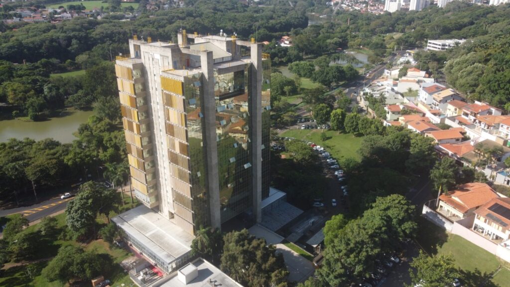 A imagem mostra uma vista aérea de um prédio alto cercado por árvores e casas. O prédio é de múltiplos andares, com uma fachada de vidro e metal, e parece ser um edifício comercial ou de escritórios. Ele está localizado em uma área arborizada, com um lago visível ao fundo. Há um estacionamento próximo ao prédio, e as casas ao redor parecem ser residenciais. A imagem é bem iluminada, sugerindo que foi tirada durante o dia. A área parece ser uma parte suburbana de uma cidade, com uma mistura de espaços verdes e desenvolvimento urbano. As árvores são densas e verdes, indicando uma área com boa vegetação. A imagem também mostra ruas e estradas, sugerindo uma infraestrutura de transporte. No geral, a imagem oferece uma visão geral de um ambiente urbano integrado à natureza.