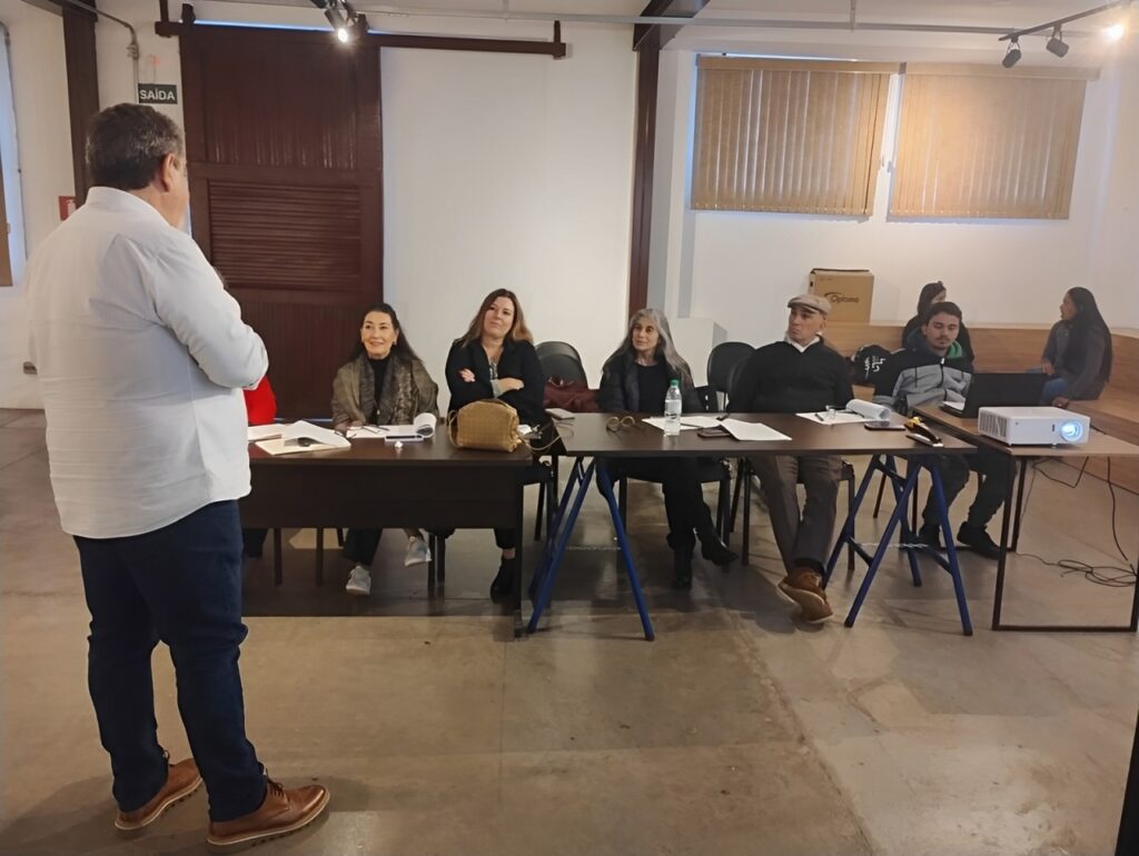 A imagem mostra uma sala de reuniões com uma pessoa de pé, de costas para a câmera, aparentemente apresentando algo a um grupo de pessoas sentadas em uma mesa. A pessoa que está de pé usa uma camisa branca e calças azuis escuras. A mesa tem vários papéis, canetas e um projetor. Há seis pessoas sentadas na mesa, três mulheres e três homens, todas com expressões atentas. As paredes são claras e há janelas com persianas. No fundo, há outras pessoas, aparentemente observando ou esperando. A atmosfera parece formal e profissional, possivelmente uma reunião de trabalho ou apresentação. A placa "SAÍDA" indica uma porta de saída.
