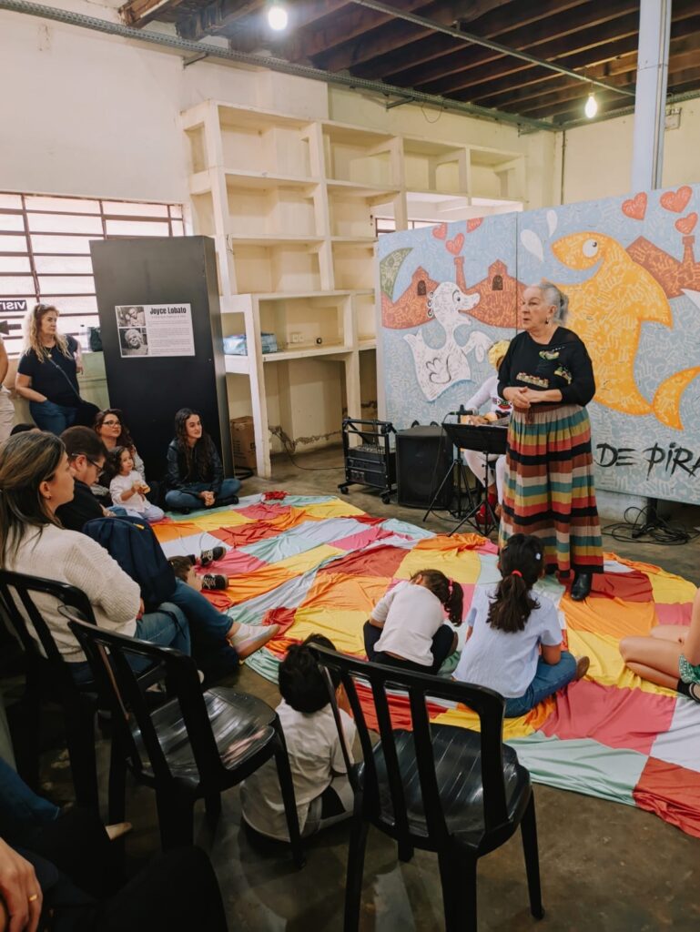 A imagem mostra um grupo de pessoas, incluindo adultos e crianças, reunidas em uma sala com um piso de concreto. No centro da sala, há uma mulher mais velha em pé, vestida com uma blusa preta e uma saia longa e colorida, que parece estar falando ou contando uma história. Ela está de frente para um mural vibrante com desenhos de animais e corações. As pessoas estão sentadas em um grande tecido colorido no chão, que parece ser um tapete ou uma área de atividade. Algumas pessoas estão sentadas em cadeiras pretas. Há uma estante vazia e alta ao fundo, e uma janela com luz natural entrando. Parece ser um evento cultural ou uma atividade para crianças e adultos, possivelmente em um centro comunitário ou espaço de arte.