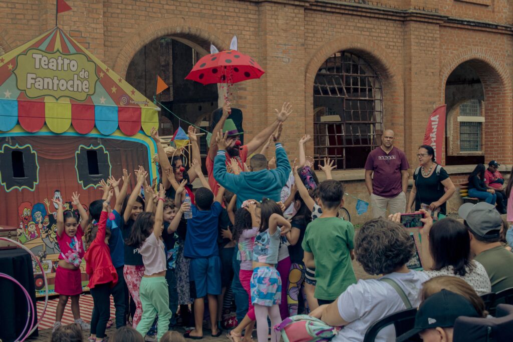 A imagem mostra um grupo de crianças animadas com as mãos levantadas em frente a uma tenda de fantoches colorida. Um palhaço com nariz vermelho e chapéu verde está no centro, segurando um guarda-chuva vermelho com bolinhas pretas, possivelmente uma joaninha. Atrás das crianças, há uma estrutura de tijolos com arcos e janelas grades. Duas pessoas adultas observam a cena, e outras pessoas estão sentadas ao fundo. A atmosfera geral é de diversão e entretenimento infantil.