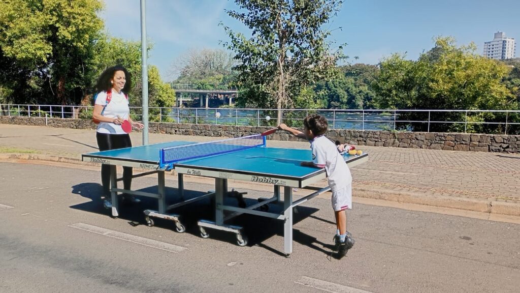 A imagem mostra uma mulher e uma criança jogando tênis de mesa ao ar livre em um dia ensolarado. Eles estão em uma área pavimentada perto de um rio, com árvores e vegetação ao fundo. A mesa de tênis de mesa é azul e branca, e ambos os jogadores estão usando roupas leves. A mulher tem cabelo escuro e comprido e está usando uma camiseta branca e calças pretas. A criança tem cabelo curto e escuro e está usando uma camiseta branca e shorts brancos. Ambos parecem estar se divertindo, e a imagem transmite um sentimento de alegria e atividade ao ar livre. O fundo inclui um rio calmo, uma ponte no fundo, e árvores e vegetação exuberante ao redor. O dia é brilhante e ensolarado, e a iluminação da imagem é natural e bem distribuída. A imagem é nítida e bem focada, e os detalhes são facilmente visíveis.