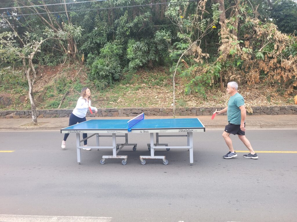A imagem mostra duas pessoas jogando tênis de mesa em uma rua. Uma mulher está de pé no lado esquerdo da mesa e um homem está de pé no lado direito. A mesa de tênis de mesa é azul e branca e está posicionada no meio da rua. Há árvores e arbustos ao fundo. O asfalto da rua é cinza escuro. A mulher está usando calças pretas e uma camisa branca. O homem está usando shorts pretos e uma camisa verde-escura. Ambos parecem estar se divertindo e jogando com entusiasmo. O cenário sugere um ambiente informal e descontraído, possivelmente uma partida amistosa entre amigos ou familiares. A iluminação na imagem indica que provavelmente é dia.