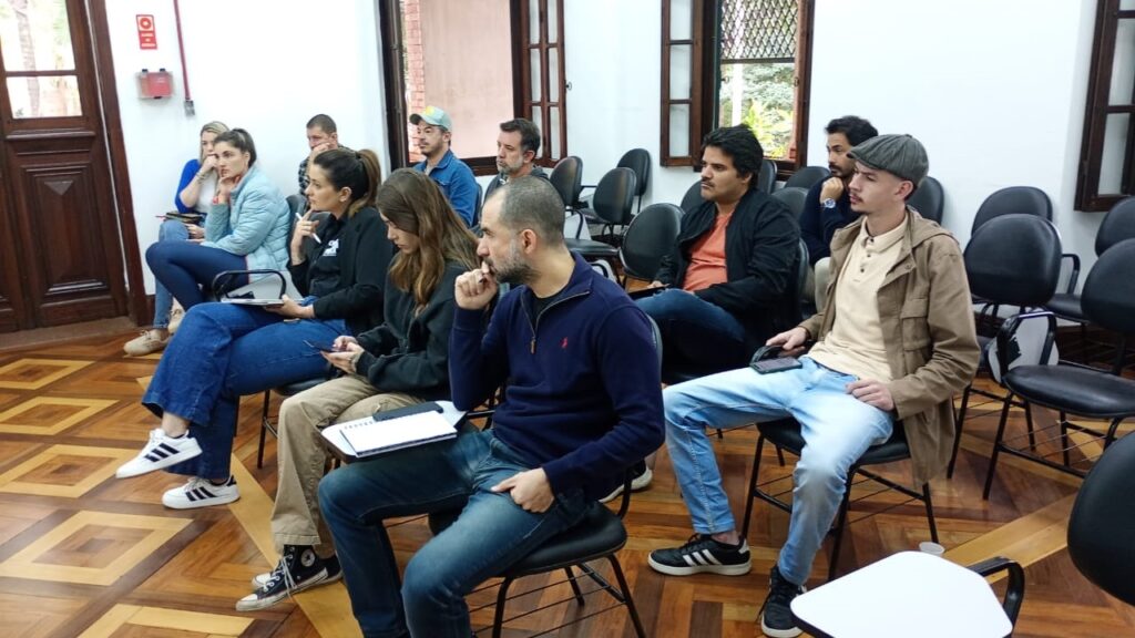 A imagem mostra um grupo de pessoas sentadas em cadeiras em uma sala. Há aproximadamente 12 pessoas, a maioria parece ser adultos jovens. Estão sentadas em fileiras, algumas em cadeiras individuais e outras em grupos de duas ou três. A sala parece ser uma sala de aula ou sala de reuniões, com cadeiras pretas e um piso de madeira. As pessoas parecem estar em uma reunião ou apresentação, algumas estão olhando para frente, enquanto outras estão conversando entre si. Há um quadro branco na parte inferior direita da imagem. A atmosfera parece formal, mas descontraída. A iluminação é natural, vindas de janelas grandes que se veem ao fundo.