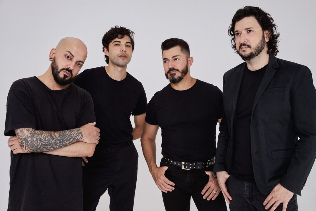 A imagem mostra quatro homens vestidos de preto, posados contra um fundo branco. O homem mais à esquerda é careca e tem uma tatuagem no braço. Ele está com os braços cruzados. O homem ao seu lado tem cabelo castanho escuro e está com uma camiseta preta. O terceiro homem tem cabelo castanho escuro e barba, e está com uma camiseta preta e cinto com detalhes metálicos. O homem mais à direita está usando um blazer preto sobre uma camiseta preta e está com os braços cruzados. Todos parecem estar olhando para a câmera. A imagem parece ser um retrato promocional de uma banda ou grupo musical.
