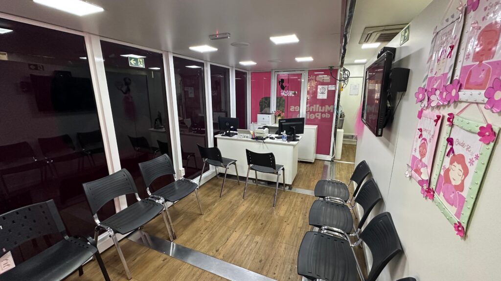 A imagem mostra uma pequena sala de espera, possivelmente de um consultório médico ou clínica. A sala é decorada em tons de rosa e branco, com alguns quadros na parede com mensagens de empoderamento feminino. Há uma recepção com dois computadores e algumas cadeiras para os pacientes esperarem. O chão é de madeira e as paredes são brancas. A iluminação é artificial, provavelmente de lâmpadas fluorescentes no teto. A sala parece limpa e organizada. Há também uma televisão na parede. No geral, a imagem transmite uma sensação de calma e profissionalismo.