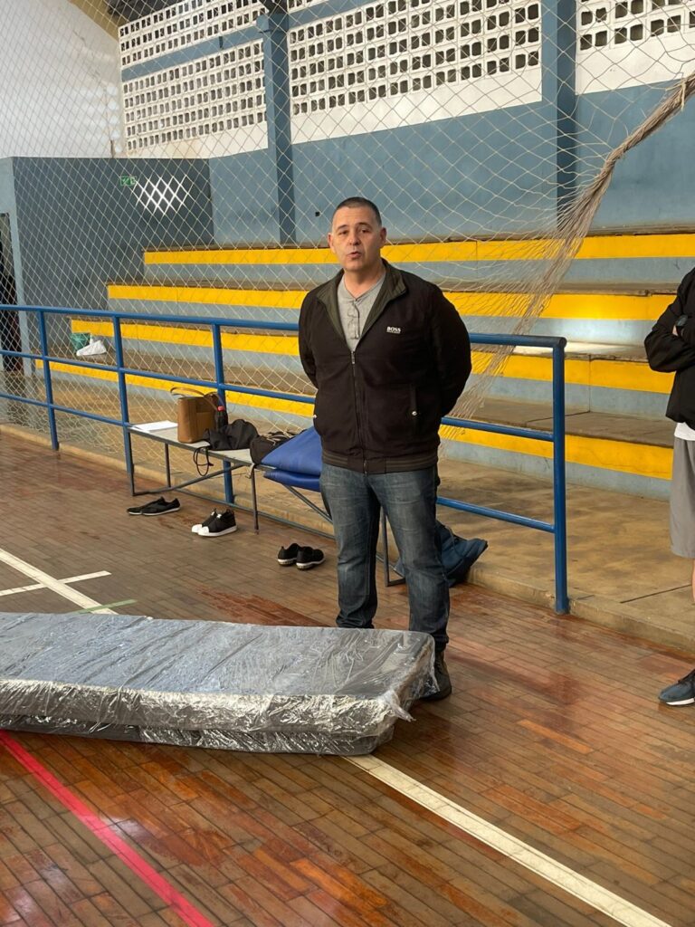 A imagem mostra um homem em pé em um ginásio, com um colchão embalado em plástico no chão à sua frente. O homem está vestindo uma jaqueta preta, uma camisa cinza e jeans. Ele tem as mãos atrás das costas e está olhando para a câmera. Atrás dele, há um conjunto de arquibancadas azuis e amarelas e uma rede de voleibol. Há também uma mesa com uma bolsa e alguns sapatos em um dos degraus da arquibancada. O chão do ginásio é de madeira e tem algumas marcas brancas e vermelhas.