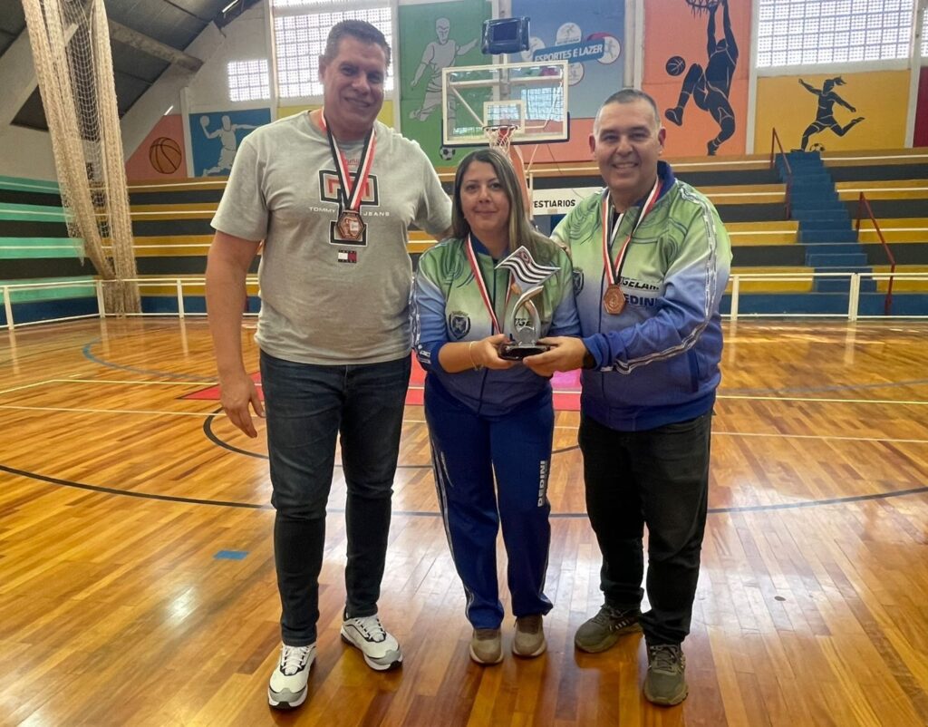 Na imagem, três pessoas estão em uma quadra de esportes interna com piso de madeira. Uma mulher no centro segura um troféu com a bandeira de São Paulo. Ela está usando calças de moletom azuis e um agasalho com detalhes em verde e azul. À esquerda dela, um homem alto, vestindo uma camiseta cinza da Tommy Hilfiger e jeans escuros, usa uma medalha no pescoço. À direita da mulher, um homem com um agasalho verde e azul similar ao dela, também com uma medalha, sorri para a câmera. O fundo mostra uma quadra de basquete com cestas e arquibancadas azuis e amarelas. As paredes atrás das arquibancadas são decoradas com murais coloridos que retratam atletas em movimento, como jogadores de basquete e futebol. A iluminação geral sugere um ambiente esportivo interno.
