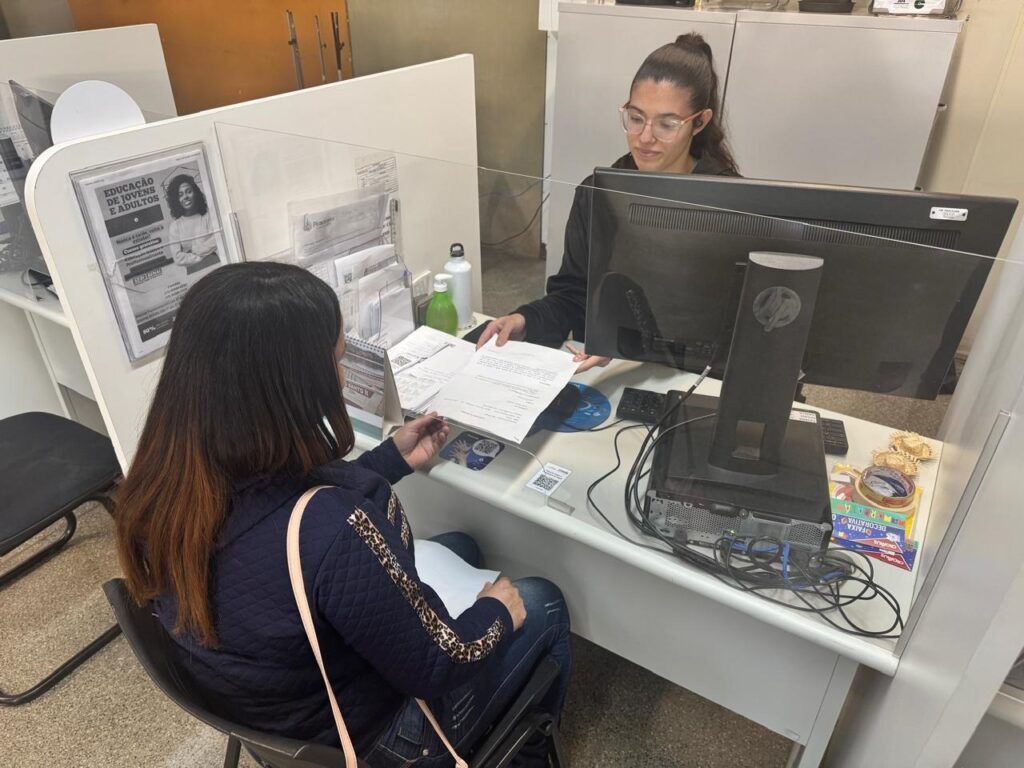 A imagem mostra duas mulheres em um balcão de atendimento, separadas por um protetor de acrílico. Uma das mulheres está sentada em uma cadeira, enquanto a outra está atrás de um computador. Há vários papéis e documentos na mesa, e um cartaz na parede com informações sobre educação para jovens e adultos. O ambiente parece ser um escritório público ou uma instituição de ensino. A atmosfera é calma e profissional.