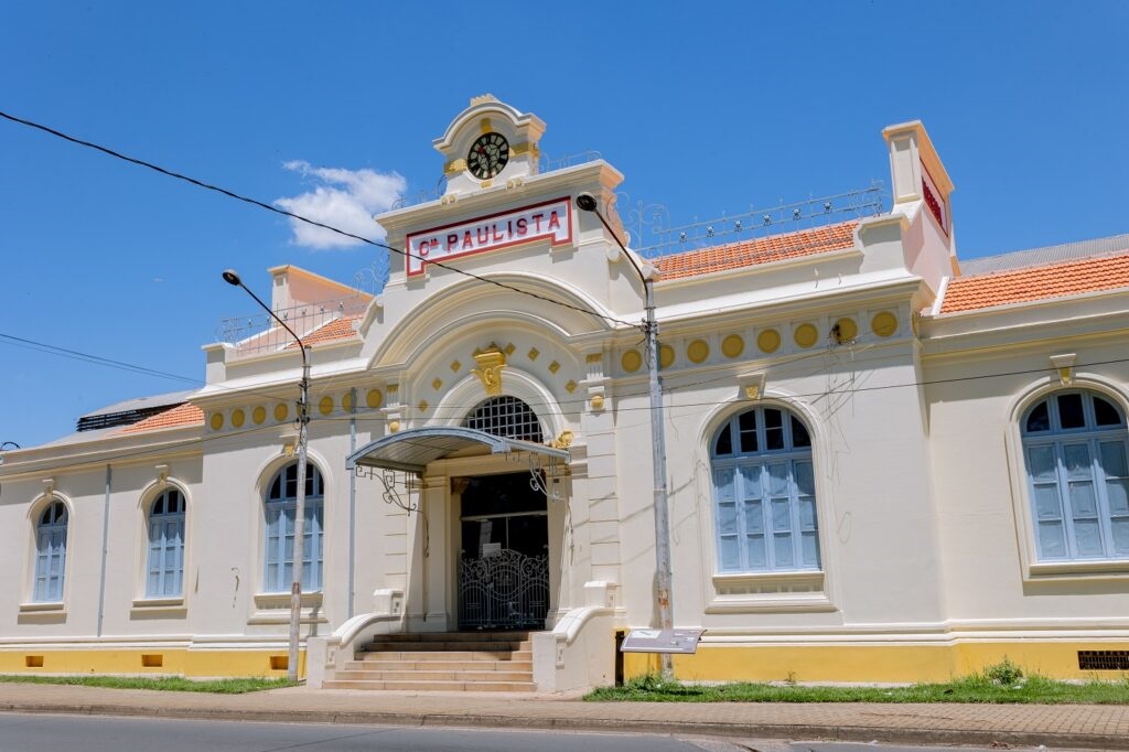 A imagem mostra uma construção de estilo arquitetônico clássico, possivelmente uma estação de trem ou edifício público. O prédio é de cor creme, com detalhes em amarelo dourado ao redor das janelas e na entrada principal. As janelas são grandes e arqueadas, com vidros azuis claros. A entrada principal possui uma marquise e uma escada de acesso. Há um relógio na parte superior do edifício, e um letreiro com a inscrição "Cª PAULISTA" indica o nome do local. A fachada é limpa e bem conservada, com um telhado de telhas vermelhas. O céu é azul e sem nuvens, indicando um dia ensolarado. A rua em frente ao prédio parece tranquila, sem muitos veículos ou pessoas. A imagem sugere um ambiente histórico e bem preservado.