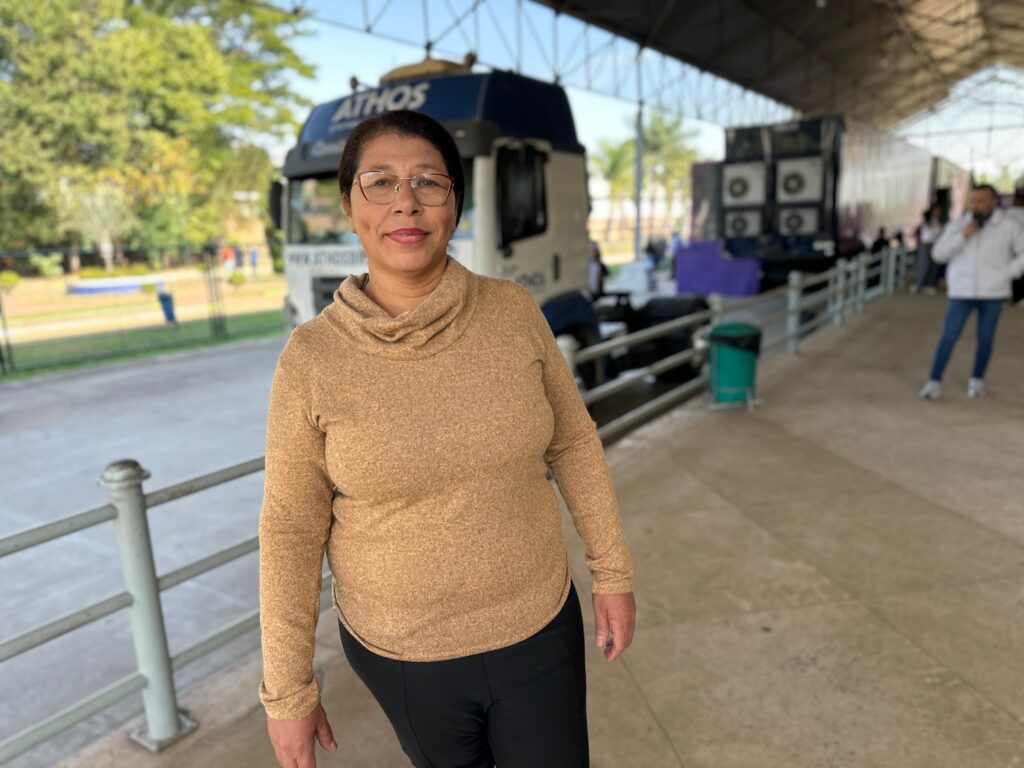 A imagem mostra uma mulher de meia-idade com óculos, vestindo um suéter bege e calças pretas, caminhando em direção à câmera. Ela parece calma e sorridente. Ao fundo, há caminhões grandes e um ambiente que parece ser um pátio ou terminal de transporte. O cenário sugere um local de trabalho ou transporte, com outras pessoas visíveis ao fundo, mas fora de foco. A mulher é o foco principal da imagem, e sua expressão facial e postura transmitem uma sensação de tranquilidade e confiança. O ambiente é iluminado por luz natural, criando uma atmosfera clara e diurna.