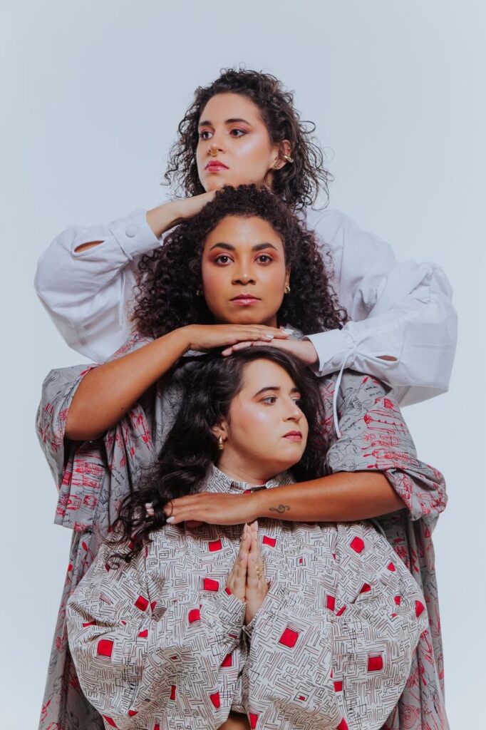 A imagem mostra três mulheres com cabelos cacheados em close-up, empilhadas umas sobre as outras. A mulher do topo, com um nariz piercing e um brinco de argola, está com a mão na cabeça da mulher do meio. A mulher do meio, com um tom de pele mais escuro e maquiagem nos olhos, está com as mãos na cabeça da mulher de baixo. A mulher de baixo, com uma tatuagem no braço e um anel em forma de flor, está com as mãos juntas em posição de oração. Todas as mulheres estão vestindo roupas com padrões distintos. A mulher do topo usa uma camisa branca com mangas bufantes, enquanto as outras duas usam camisas com estampas geométricas em tons de cinza, vermelho e preto. O fundo é branco e liso, destacando as mulheres.