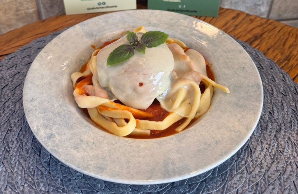 A imagem mostra um prato de massa com um molho vermelho e uma grande porção de queijo derretido por cima, decorado com folhas de manjericão. A massa parece ser fresca e artesanal, com uma textura ligeiramente grossa e irregular. O molho vermelho, que cobre a massa, tem uma cor vibrante e parece ser feito com tomates frescos. O queijo derretido, que cobre o topo do prato, é branco e cremoso, com alguns pontos dourados de cozimento. As folhas de manjericão, de cor verde escura, adicionam um toque de frescor e cor ao prato. O prato está colocado sobre uma toalha de mesa cinza com uma textura entrelaçada, que contrasta com a textura lisa do prato de cerâmica cinza claro com manchas. Ao fundo, desfocados, aparecem dois pequenos cartões verdes com texto em branco, sugerindo que o prato faz parte de um menu ou de uma oferta especial de um restaurante ou delicatessen. A iluminação é suave e natural, realçando as cores e texturas da comida.