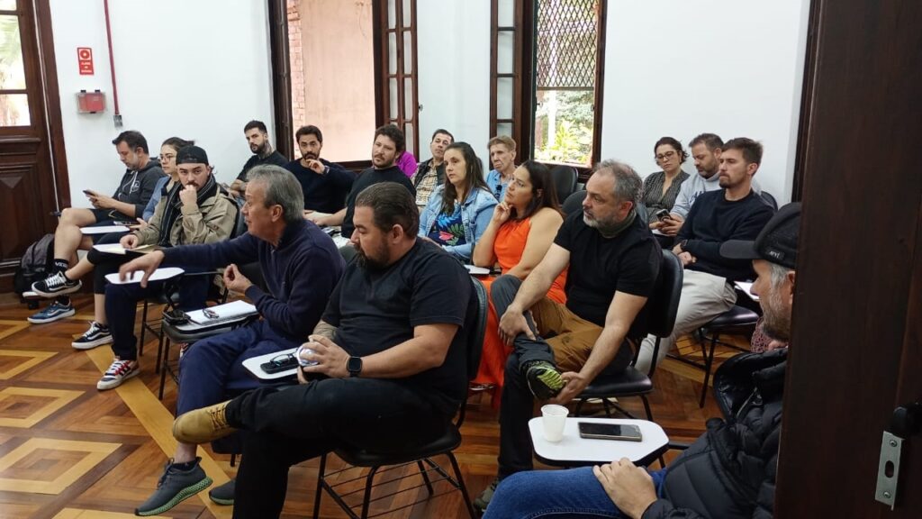 A imagem mostra um grupo de pessoas sentadas em cadeiras em uma sala. A maioria das pessoas parece estar envolvida em uma discussão ou apresentação. A sala tem um piso de madeira e paredes claras. As pessoas estão sentadas em fileiras, e há uma mistura de homens e mulheres. Algumas pessoas estão usando roupas formais, enquanto outras estão usando roupas mais casuais. Há uma mesa na frente de algumas das pessoas, e alguns papéis e canetas estão sobre ela. A imagem parece ter sido tirada em um ambiente acadêmico ou de negócios.