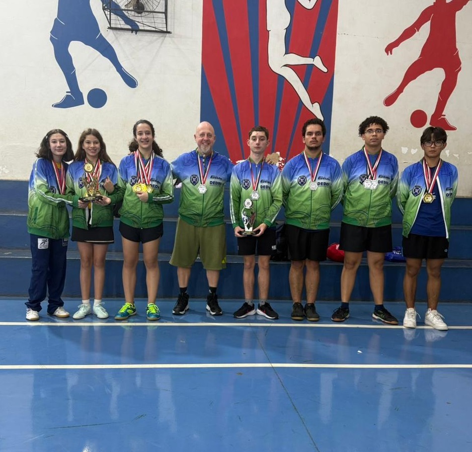 A imagem mostra um grupo de oito pessoas, incluindo três mulheres jovens e cinco homens jovens, posando para uma foto em uma quadra esportiva. Todos estão vestindo jaquetas esportivas com as cores verde e azul, e a maioria usa medalhas no pescoço. As mulheres mais à esquerda e a terceira mulher da esquerda para a direita estão segurando troféus e medalhas de ouro. O homem no centro, que parece ser um treinador ou supervisor, também está usando uma medalha. Os outros homens jovens também estão usando medalhas, a maioria de prata. Ao fundo, há uma parede com desenhos de silhuetas de jogadores de futebol e um logotipo com listras vermelhas e azuis. O piso da quadra é azul.