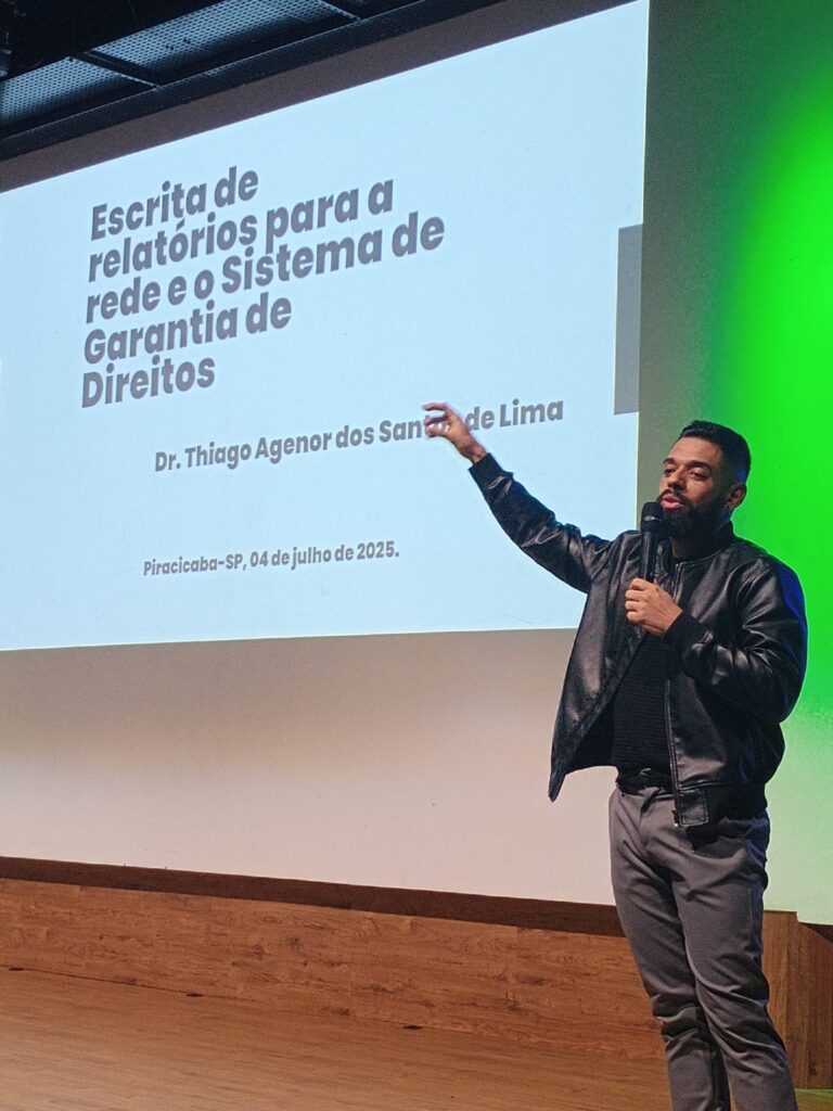 A imagem mostra um homem de pele morena, com barba e bigode, vestindo uma jaqueta de couro preta e calças cinza, dando uma apresentação. Ele está de pé em um palco, com uma tela de projeção atrás dele exibindo o título de sua apresentação: "Escrita de relatórios para a rede e o Sistema de Garantia de Direitos", seguido pelo seu nome, "Dr. Thiago Agenor dos Santos de Lima", e a data: "Piracicaba-SP, 04 de julho de 2025". Ele segura um microfone e aponta para a tela com a mão direita. A iluminação do palco é suave, com uma parede verde visível à direita da tela. O chão é de madeira escura. A imagem sugere uma apresentação formal em um evento ou conferência.