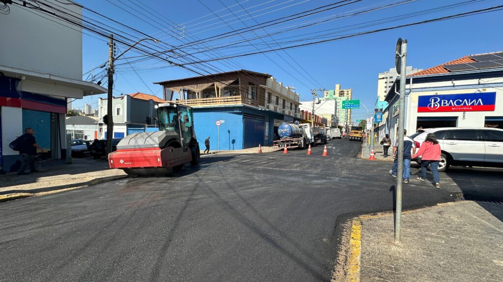 A imagem mostra uma rua em processo de asfaltamento. Uma compactadora de asfalto vermelha está no primeiro plano, recém-passada sobre o asfalto fresco. Há um caminhão tanque azul estacionado mais adiante, aparentemente fornecendo o asfalto. Cones de segurança laranja marcam o perímetro da área de trabalho. No fundo, há prédios residenciais e comerciais de um e dois andares, com fiação elétrica aérea visível. Várias pessoas estão presentes na cena, algumas observando o trabalho e outras andando pela rua. O céu está claro e azul. O asfalto parece recém-aplicado e ainda úmido em alguns pontos. A cena sugere uma obra pública de manutenção ou melhoria viária em andamento em uma área urbana.