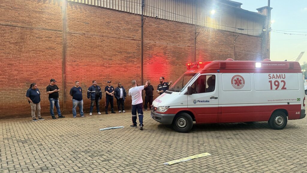 Um grupo de pessoas, algumas em uniforme e outras em roupas casuais, está reunido em uma área externa com uma parede de tijolos ao fundo. Uma ambulância, identificada pelas marcações "SAMU 192" e pela combinação de cores vermelha e branca, está estacionada no lado direito da imagem. A ambulância tem suas luzes de emergência ativadas, emitindo um brilho vermelho sobre a cena. Algumas pessoas parecem estar ouvindo um indivíduo vestido com uma camisa branca que está gesticulando. O chão é pavimentado com tijolos interligados. A impressão geral é de um briefing ou sessão de treinamento envolvendo pessoal médico de emergência.