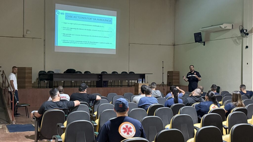 A imagem mostra uma sala de treinamento ou reunião, onde um homem vestindo um uniforme escuro, possivelmente um profissional de saúde ou socorrista, está em pé e falando para uma audiência sentada em cadeiras. Ele está de frente para uma tela grande onde se lê "CABE AO 'CONDUTOR' DA AMBULÂNCIA" e uma lista de responsabilidades. A audiência, composta por homens e mulheres, está sentada em cadeiras dispostas em fileiras, voltadas para a tela e o palestrante. Muitos dos presentes também vestem uniformes escuros, alguns com o logo do SAMU. O ambiente parece ser um auditório ou sala de aula, com iluminação artificial e um ventilador de teto visível. Há também uma mesa com garrafas de água à frente da audiência. Um homem, também de uniforme, está em pé no lado esquerdo da sala, perto da entrada.