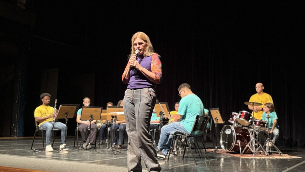 A imagem mostra uma mulher de meia-idade, com cabelos loiros e um microfone na mão, falando para uma plateia. Ela está vestindo uma blusa roxa e calças jeans. Ao fundo, há uma banda composta por vários jovens tocando instrumentos musicais, como clarinetes, saxofones e bateria. Os jovens estão sentados em cadeiras, e a banda parece estar em um palco. A iluminação é focada na mulher que fala, mas a banda também está visível e bem iluminada. A atmosfera parece ser de um evento ou apresentação musical. A mulher parece estar conduzindo ou apresentando a apresentação da banda.